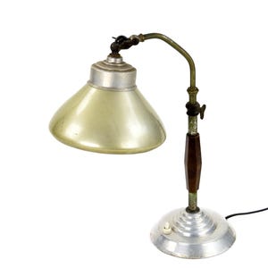 Puede incluir: Lámpara de escritorio vintage con base y pantalla plateadas. La lámpara tiene un brazo curvo y un acento de madera marrón oscuro en el tallo. Un interruptor de encendido/apagado está ubicado en la base. La lámpara está diseñada para uso en escritorio.