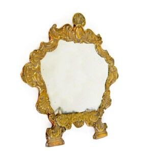 May include: Ornate, antique-style vanity mirror with a gold-colored frame. The mirror has an oval shape and is supported by two small feet. The frame features intricate scrollwork and a shell-shaped accent at the top.