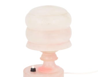 May include: A small, pink table lamp with a rounded, stacked shade and a pedestal base. The lamp has a small on/off switch and a power cord. The lamp is made of a translucent material, giving it a soft, diffused glow when lit.
