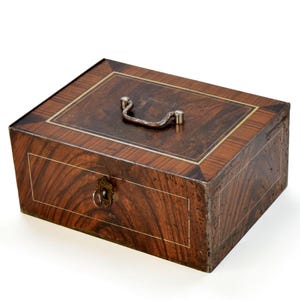 Könnte beinhalten: Eine rechteckige Holzbox mit dunkelbrauner Holzmaserung und hellen Einlegearbeiten an den Rändern. Die Oberseite ist mit einem silberfarbenen Metallgriff versehen, und an der Vorderseite befindet sich ein Schlüsselloch mit einem silbernen Schlüssel.