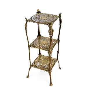 Puede incluir: Un soporte de latón ornamentado de tres niveles con intrincados diseños florales y de volutas. Los estantes redondos están conectados por soportes delgados y decorativos. El soporte tiene una estética vintage y es adecuado para exhibir objetos decorativos.