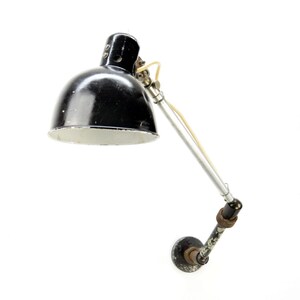 Puede incluir: Lámpara de escritorio de pared negra y plateada con una pantalla redondeada. La lámpara tiene un brazo ajustable y una base circular. El interior de la pantalla es blanco y un cable amarillo sale de la lámpara.