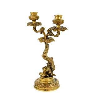 May include: A gold-colored, ornate candelabra with two candle holders. The base is round with floral detailing, and the stem is shaped like a dolphin. The candle holders are surrounded by decorative floral accents.