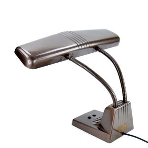Könnte beinhalten: Eine vintage braune Schreibtischlampe mit rechteckigem Schirm und flexiblen Schwanenhalsarmen. Die Lampe hat einen rechteckigen Sockel und ein Netzkabel. Der Schirm hat drei horizontale Linien.
