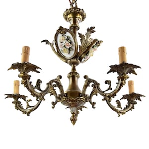 May include: Ornate brass chandelier with five candle holders. The fixture features decorative floral accents and a central oval porcelain panel with painted roses. The candle holders have a leaf design.