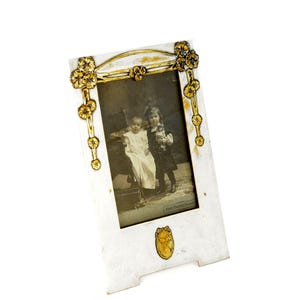 Op de afbeelding: Wit en goud Art Nouveau fotolijst met een zwart-wit foto van twee kinderen. Het frame heeft gouden bloemen en gebogen accenten. De rechthoekige foto heeft onderaan de tekst "Hans Friderich Monogram".
