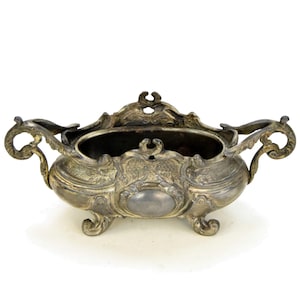 May include: Ornate silver-toned metal bowl with decorative handles and feet. The bowl features detailed scrollwork and an oval embellishment on the front. The interior of the bowl is dark, contrasting with the bright exterior. A vintage decorative piece.