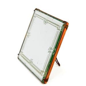 Puede incluir: Un espejo cuadrado vintage con un borde decorativo grabado. El espejo está enmarcado en un marco de madera con un borde de metal color cobre. El marco tiene un soporte incorporado. El espejo está en ángulo, mostrando su superficie reflectante.
