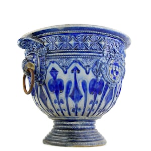May include: A large, ornate blue and white ceramic planter with intricate relief patterns. The planter features a detailed design with floral and geometric motifs, a pedestal base, and a rusty metal ring handle. The overall style is antique.