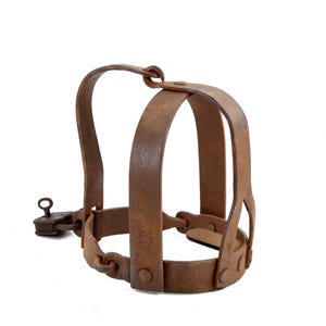 May include: A rusty brown metal muzzle with a hinged lock and a curved handle. The muzzle has a cage-like design with vertical and horizontal bars, and is riveted together. The object is isolated on a white background.