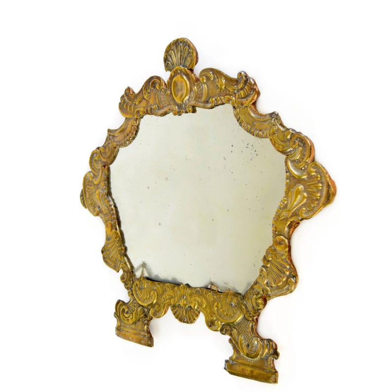May include: Ornate antique mirror with a gold-colored, decorative frame. The mirror has an irregular shape and is supported by two small feet. The frame features intricate scrollwork and a shell-like design at the top.