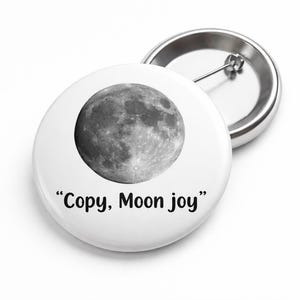 May include: White button with a grayscale image of the moon and the text "Copy, Moon joy". A second button with a pin back is visible in the background. The buttons are round and have a shiny metal finish.