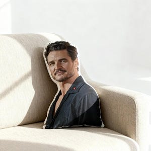 May include: A decorative pillow featuring a printed image of a man in a dark blue jacket. The pillow is shaped to the outline of the man's head and shoulders. The pillow is resting on a beige sofa.