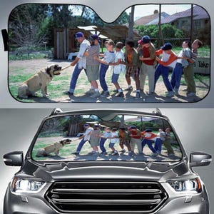 May include: Car windshield sunshade featuring a scene of a group of children playing tug-of-war with a large dog. The image is a still from the movie "The Sandlot". The sunshade is black with a reflective surface.