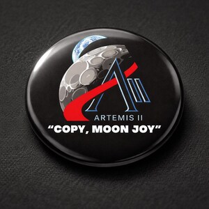 May include: A black button with the Artemis II logo, featuring a stylized moon, Earth, and the text "COPY, MOON JOY". The logo includes red and blue accents.