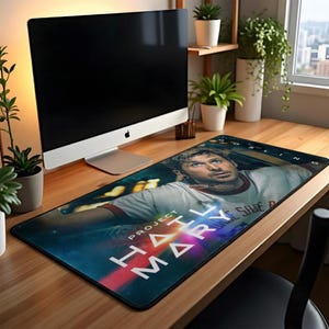 May include: A rectangular mousepad featuring a movie poster design with the text "PROJECT HAIL MARY" and a portrait of a man. The mousepad is on a wooden desk with a computer and plants.
