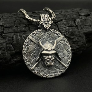 May include: A silver-colored pendant necklace featuring a detailed samurai mask design. The round pendant has a textured background with crossed swords and a helmeted face. The necklace is displayed on a dark, textured surface.