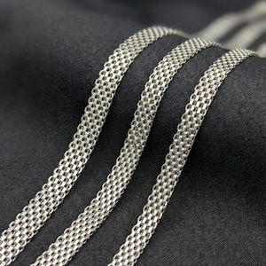 Könnte beinhalten: Nahaufnahme von drei silberfarbenen Kettenhalsketten. Die Ketten haben ein gewebtes Design mit kleinen, miteinander verbundenen Gliedern. Die Ketten werden vor einem dunklen, strukturierten Hintergrund präsentiert, der ihren metallischen Glanz hervorhebt. Die Ketten sind wahrscheinlich Schmuck.