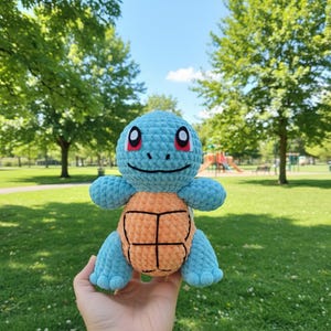 May include: A handmade crochet Squirtle plush toy. The plush features a light blue body, orange shell, and black facial details. The toy is held in front of a green park background with trees and a blue sky.
