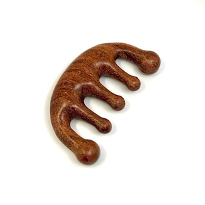 May include: A wooden comb with a curved handle and five rounded tines. The comb is made of reddish-brown wood with a visible grain pattern. The design is ergonomic and appears to be for scalp massage.