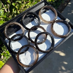 May include: A collection of seven translucent, smoky-brown circular bangles arranged in a black display tray. Each bangle features a smooth, polished surface with varying degrees of dark brown and grey tones, and some include small white tags.