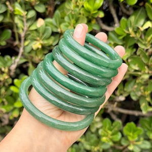 May include: A stack of jade bangles in varying sizes. The smooth, polished bracelets are a vibrant green color. The bangles are held in a hand, with a background of green foliage.