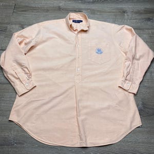 May include: A light peach-colored long-sleeve button-down shirt with a partial front placket. The shirt features a button-down collar, a single chest pocket with a blue circular embroidered logo, and buttoned cuffs. The label reads Ralph Lauren.
