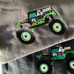 May include: Two gray sweatshirts with embroidered monster truck designs. The trucks are green with black tires and the word "GRAVE" above the cab. The design includes a skull and flames.