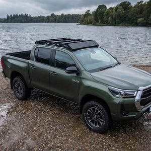 May include: An olive green Toyota Hilux pickup truck featuring a black roof rack, four doors, and dark grey alloy wheels. The vehicle is parked on a gravel shore with a large body of water and a forest visible in the background.