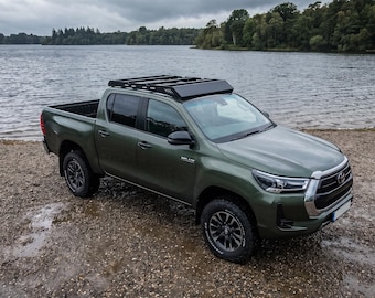 May include: An olive green Toyota Hilux pickup truck featuring a black roof rack, four doors, and dark gray alloy wheels. The vehicle is parked on a gravel shore with a large body of water and a forest visible in the background.
