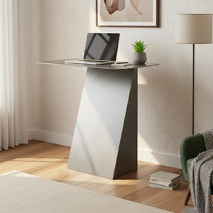 Puede incluir: Un escritorio gris moderno con una computadora portátil, una planta pequeña y un teléfono. El escritorio tiene un diseño angular único. Un sillón verde con una manta y una lámpara de pie están en el fondo. La habitación tiene una paleta de colores neutros.