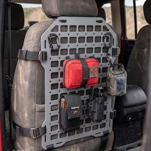Pode incluir: Um painel modular cinza para o encosto do banco de um veículo com uma grade de recortes retangulares para fixar equipamentos. O painel é preso com tiras de nylon preto e contém uma bolsa médica vermelha, um coldre de ferramentas preto e lanterna.
