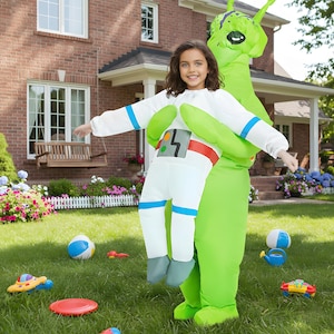 May include: A young person in a white astronaut costume is held by a person in a large green inflatable alien costume. The astronaut suit has blue and red accents. Toys are scattered on the green grass.