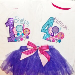 Puede incluir: Dos camisetas blancas con n&uacute;meros morados y azules, caramelos y los nombres Bailey y Addison. Un tut&uacute; de tul morado con un lazo rosa de lunares blancos.