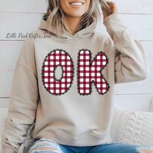以下が含まれることがあります： 赤と白のチェック柄の「OK」の文字が入ったベージュ色のパーカー。文字は黒いレースのトリムで縁取られています。パーカーにはドローストリングフードと長袖が付いています。