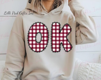 May include: Beige hoodie with the word "OK" in a red and white checkered pattern. The letters are outlined with a black lace trim. The hoodie has a drawstring hood and long sleeves.