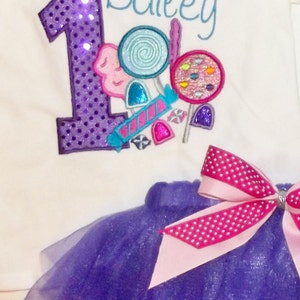 Puede incluir: Un body blanco con un n&uacute;mero uno de lentejuelas moradas y el nombre Bailey bordado en azul. El body est&aacute; decorado con dise&ntilde;os coloridos de caramelos y piruletas. Se incluye una falda de tul morada con un lazo de lunares rosa y blanco.