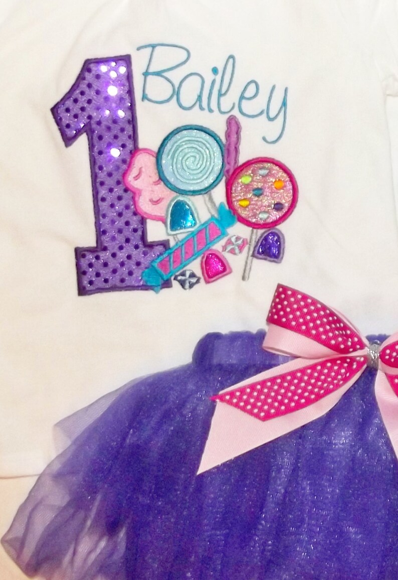 Puede incluir: Un body blanco con un n&uacute;mero uno de lentejuelas moradas y el nombre Bailey bordado en azul. El body est&aacute; decorado con dise&ntilde;os coloridos de caramelos y piruletas. Se incluye una falda de tul morada con un lazo de lunares rosa y blanco.