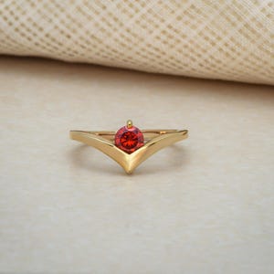 Może przedstawiać: Złoty pierścionek o kształcie litery V. Pierścionek ma okrągły, czerwony kamień szlachetny pośrodku. Pierścionek jest prezentowany na jasnej powierzchni.