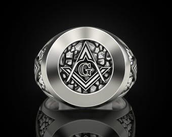 May include: A silver-toned ring with a prominent Masonic symbol at the center. The ring features a detailed design with a textured background and a polished outer band. The Masonic symbol includes a compass, square, and the letter "G".