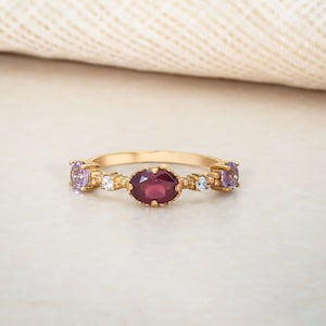 May include: A gold ring featuring a central oval-cut, deep red gemstone, flanked by two oval purple gemstones and small white accent stones. The ring's band is a polished gold color, and the gemstones are set in a delicate, ornate design.