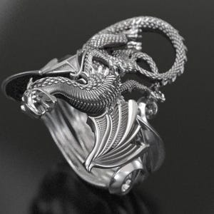 May include: A silver dragon ring. The ring showcases a detailed dragon design with wings and a coiled tail. The dragon wraps around the ring band, creating a unique and eye-catching piece of jewellery.
