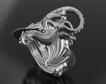 May include: A silver-colored dragon ring. The ring features a detailed dragon design with wings and a coiled tail. The dragon is wrapped around the ring band, creating a unique and eye-catching piece of jewelry.