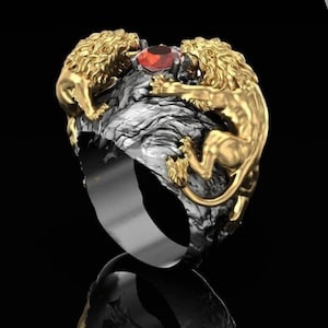 Può includere: Un anello in oro e argento con due figure di leoni dorati che fiancheggiano una gemma rossa. I leoni sono dettagliati con criniere e corpi strutturati, su una base strutturata argento e nera. L'anello ha una fascia argentata lucida.