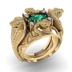 May include: A gold-coloured ring with a snake design. The ring features an emerald-green gemstone at its centre, encircled by smaller clear stones. The snakes are intricately detailed, with textured scales and small, yellow eyes. The ring is set against a white background.