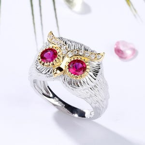 May include: A silver owl ring with gold detailing and ruby-coloured eyes. The owl's feathers are intricately designed, and the ring has a polished finish. A unique piece of jewellery.