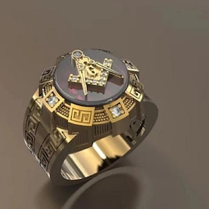 Puede incluir: Un anillo dorado con un emblema masónico con un diseño de compás y escuadra. El anillo tiene una piedra central de color rojo oscuro y está adornado con pequeñas gemas transparentes. La banda tiene un patrón geométrico detallado.