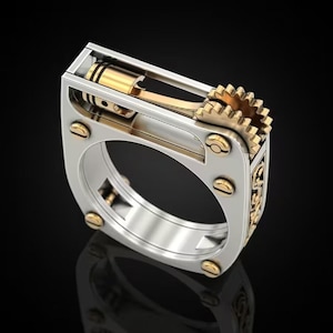 Könnte beinhalten: Ein silber- und goldfarbener Ring mit mechanischem Design. Der Ring hat einen Kolben, eine Pleuelstange und ein Zahnrad mit goldfarbenen Akzenten. Der Ring hat eine quadratische Form mit abgerundeten Kanten und goldfarbenen Nieten.