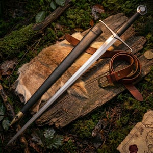 May include: A steel longsword with a black leather-wrapped grip and a straight crossguard, accompanied by a black scabbard featuring a brass chape. A brown leather belt with a brass buckle is coiled next to the sword and scabbard.