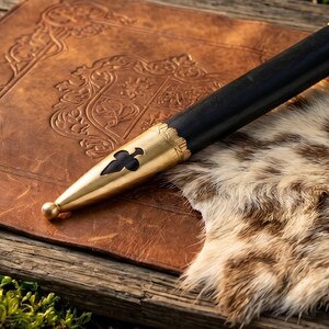 May include: A black scabbard featuring a polished brass chape at the tip. The brass fitting is adorned with a cutout in the shape of a club symbol and a small spherical finial at the very end. The scabbard body is smooth, dark, and cylindrical.